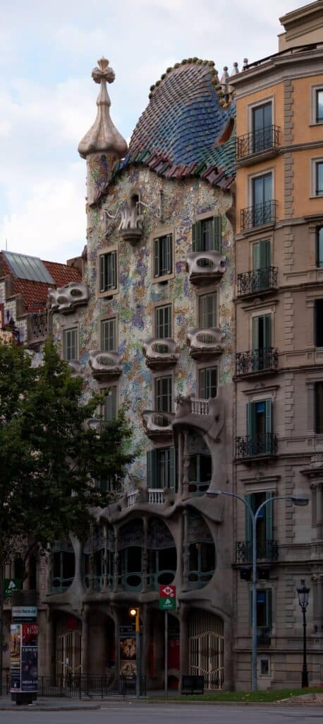 Facade of Casa Batlló on Passeig de Gràcia rising between plainer neighbours, its bone-like stone columns, mask balconies and colourful mosaic crowned by the dragon-scale roof and a cross-topped turret, trees along the pavement