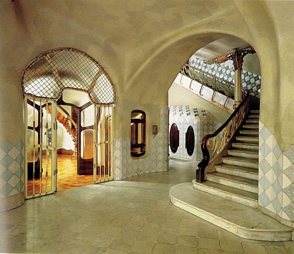 Ground floor lobby of Casa Batlló with vaulted cream walls and blue and white tiles, a wrought-iron grille doorway on the left and a stone staircase with a carved wooden handrail rising beneath an arch