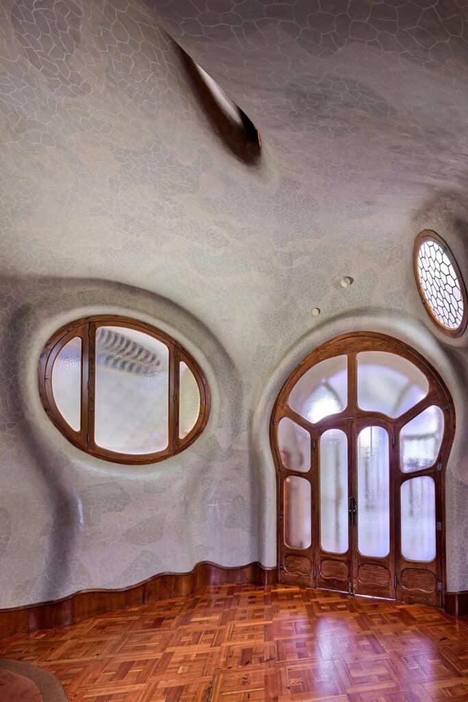 Landing of the Noble Floor at Casa Batlló with smooth curving plaster walls, an oval window and an arched oak and glass doorway, a round porthole window above and a parquet floor