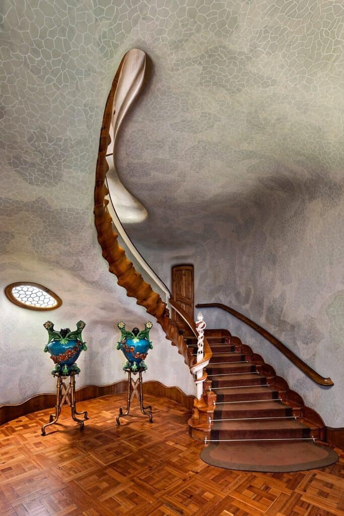 Sculptural oak staircase curling up to the Noble Floor of Casa Batlló beneath a mottled grey ceiling, with two blue-green ceramic urns on iron stands and a small oval window