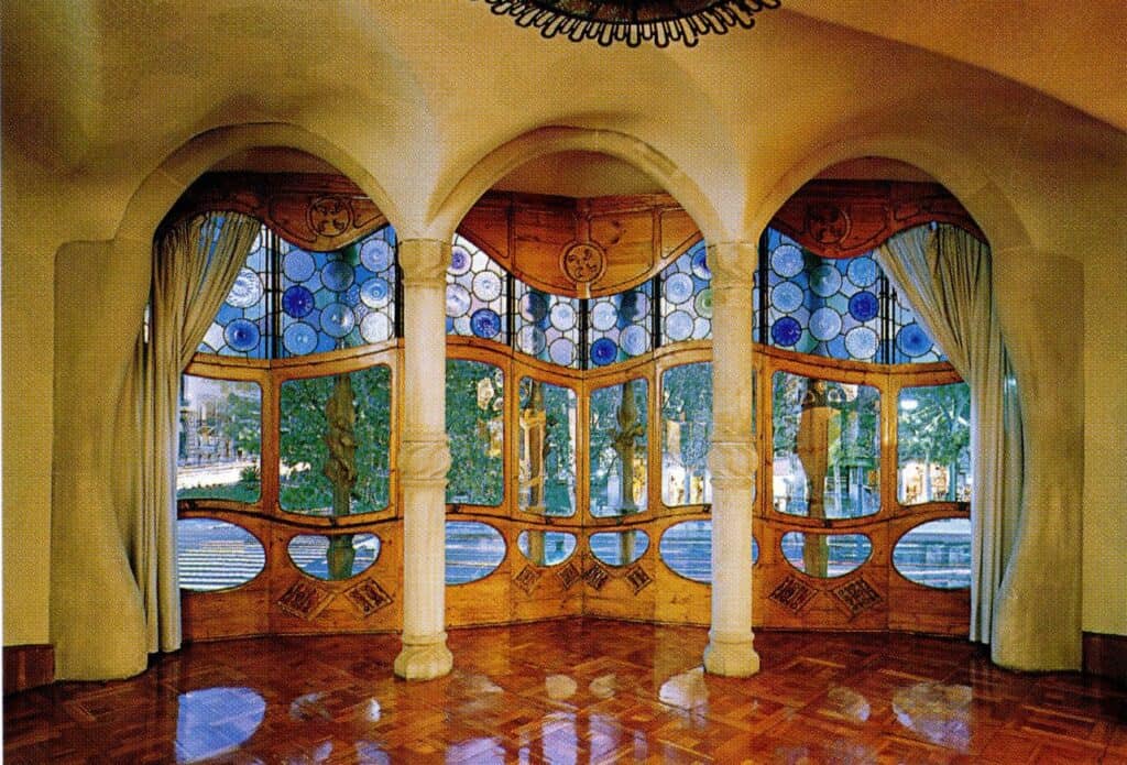 Noble Floor salon of Casa Batlló looking out through the gallery over Passeig de Gràcia, bone-like stone columns and arches framing oak windows with blue glass discs, curtains and a glossy parquet floor