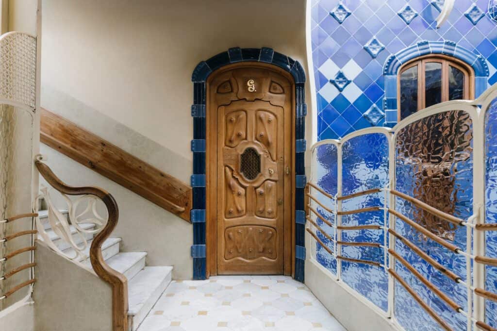 Third-floor landing of Casa Batlló with a carved oak door in a blue tile surround, rippled glass panels and wooden handrails onto the cobalt-tiled light well and a white staircase