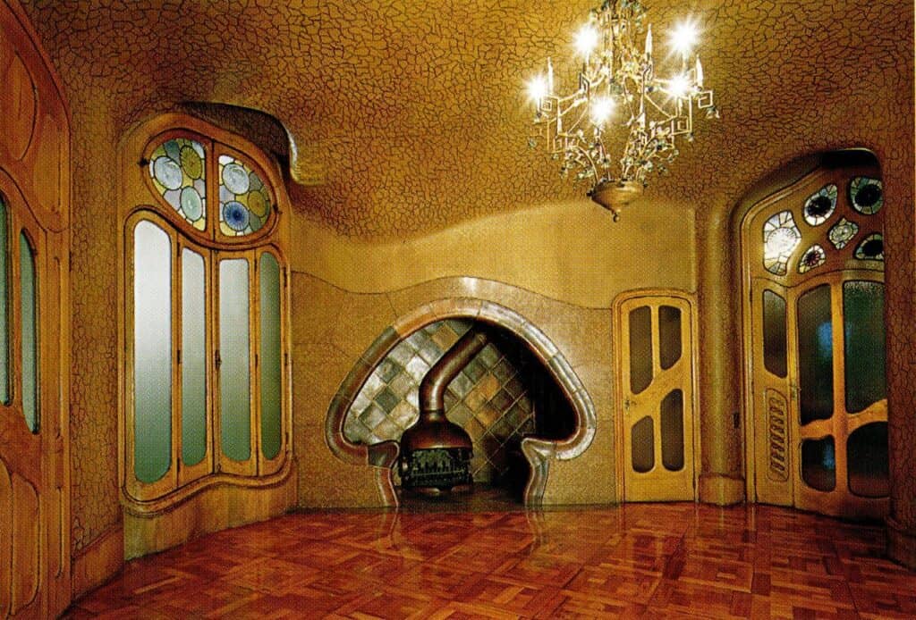 Office on the Noble Floor of Casa Batlló with a mushroom-shaped fireplace alcove set into a golden wall, stained-glass oak windows and doors, a crystal chandelier and a red parquet floor
