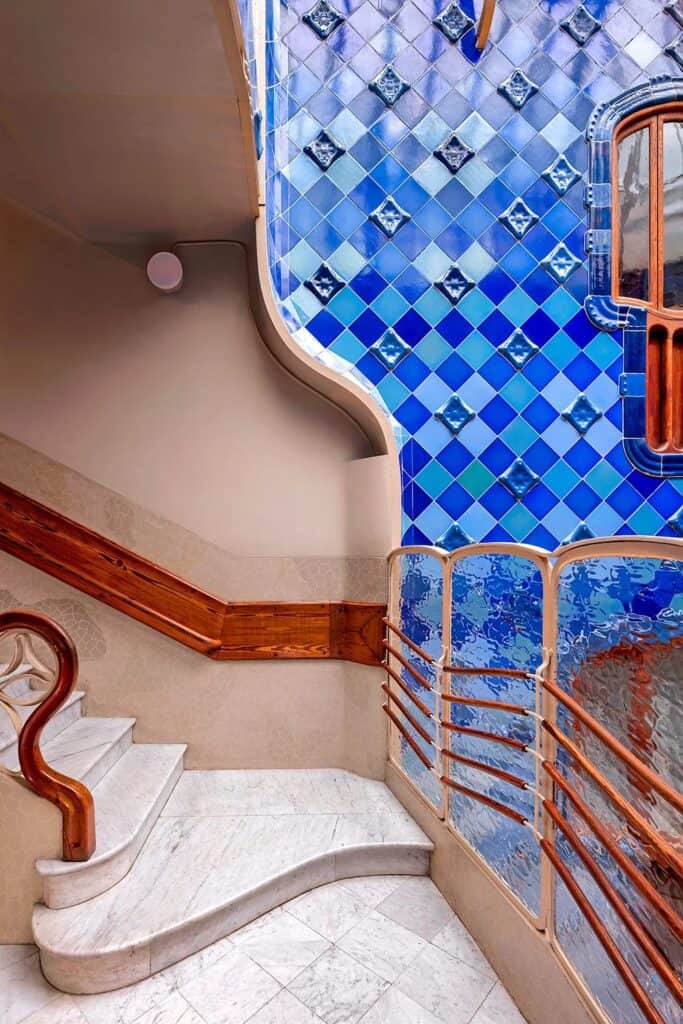 Landing beside the light well at Casa Batlló with white marble stairs, a curving oak handrail and a wavy glass balustrade before walls of blue tiles