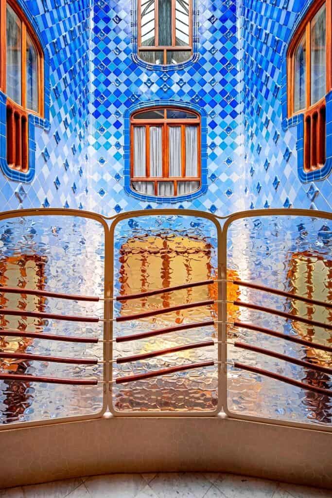 Light well of Casa Batlló lined with cobalt and pale blue diamond-patterned tiles around wooden windows, seen through a rippled glass balustrade with wooden rails