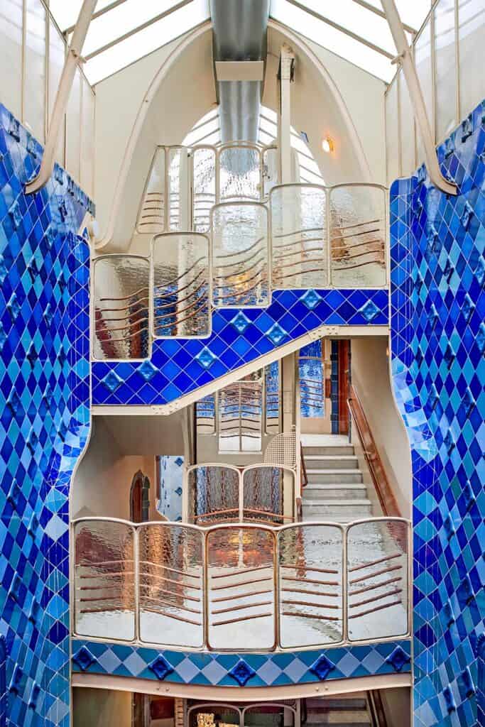 Top of the light well at Casa Batlló beneath a glazed skylight, with tiers of rippled glass balustrades, white stairs and walls of blue diamond-patterned tiles