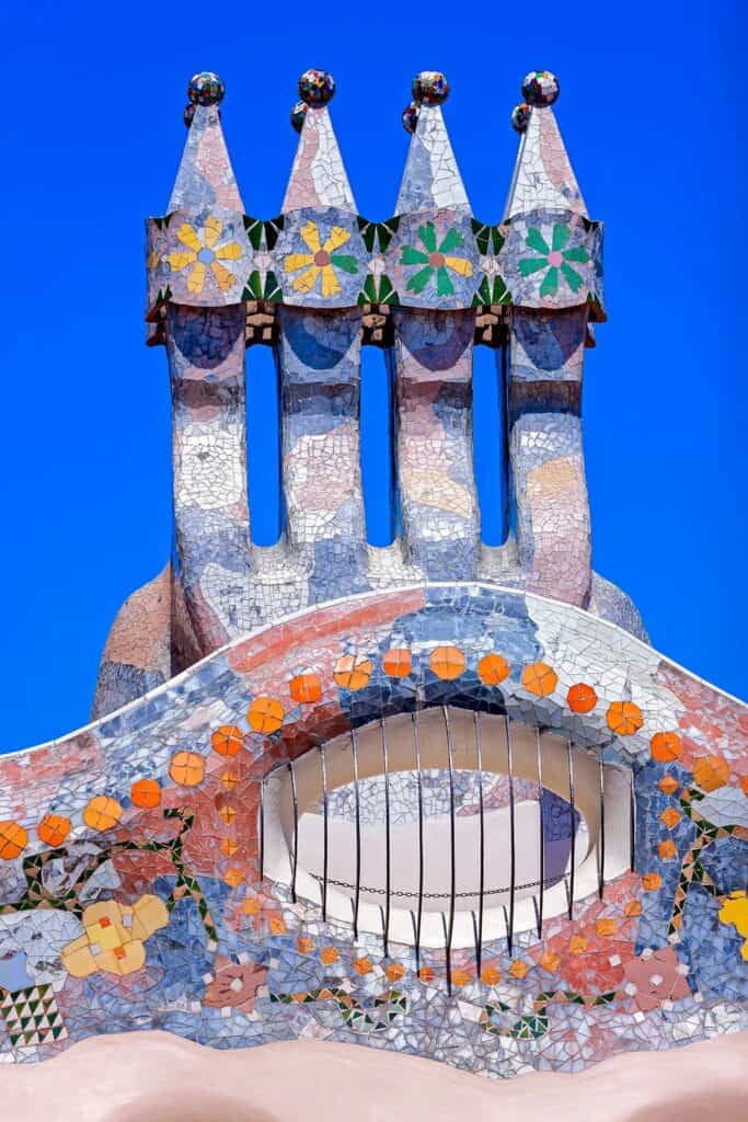 Four chimneys of Casa Batlló topped with ceramic flowers and spheres above a mosaic crown of orange and blue discs with a barred oval window, against a blue sky