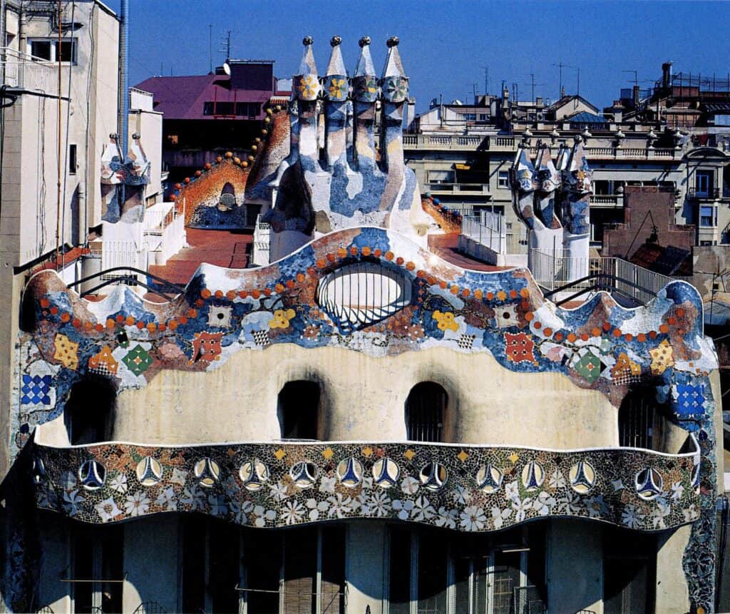 Rear facade crown of Casa Batlló with a wavy parapet of coloured ceramic discs and flowers, arched openings below and clusters of chimneys behind, over the Eixample rooftops