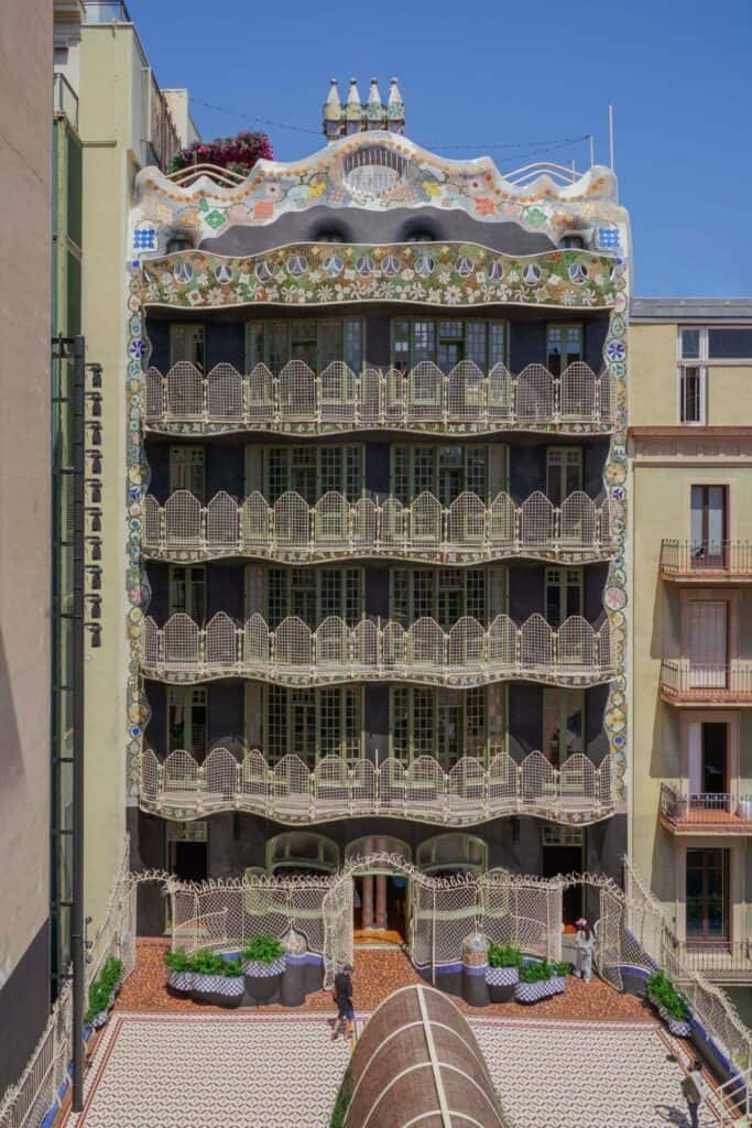 Restored rear facade of Casa Batlló above its private courtyard, with dark stucco walls, white openwork iron balconies, a mosaic crown and chimneys, and planters on the patterned courtyard floor