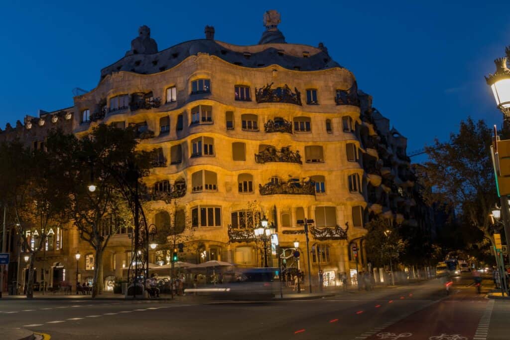 Casa Milà lit up at blue hour on the corner of Passeig de Gràcia in Barcelona, its undulating stone façade glowing gold beneath a deep blue sky, with street lamps, trees and light trails from passing traffic