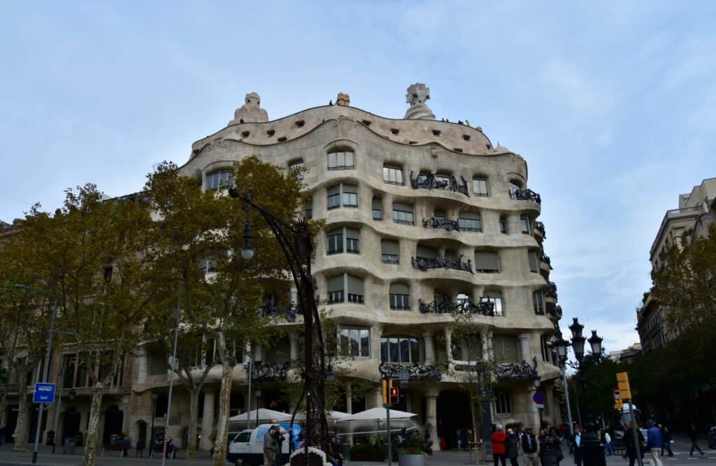 Casa Milà by day seen across Passeig de Gràcia, the rippling cream limestone façade and dark wrought-iron balconies rising behind plane trees and café umbrellas under a pale sky