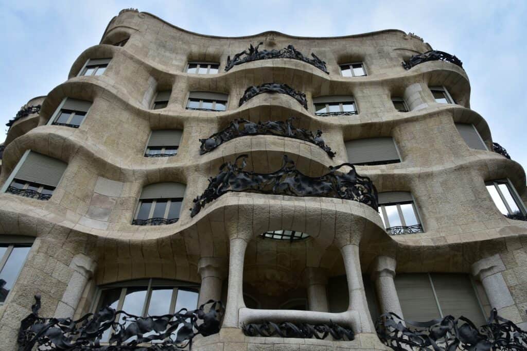 Looking up at the waving stone façade of La Pedrera, curved window openings and slender stone columns at the base, with tangled black wrought-iron balconies wrapping each floor