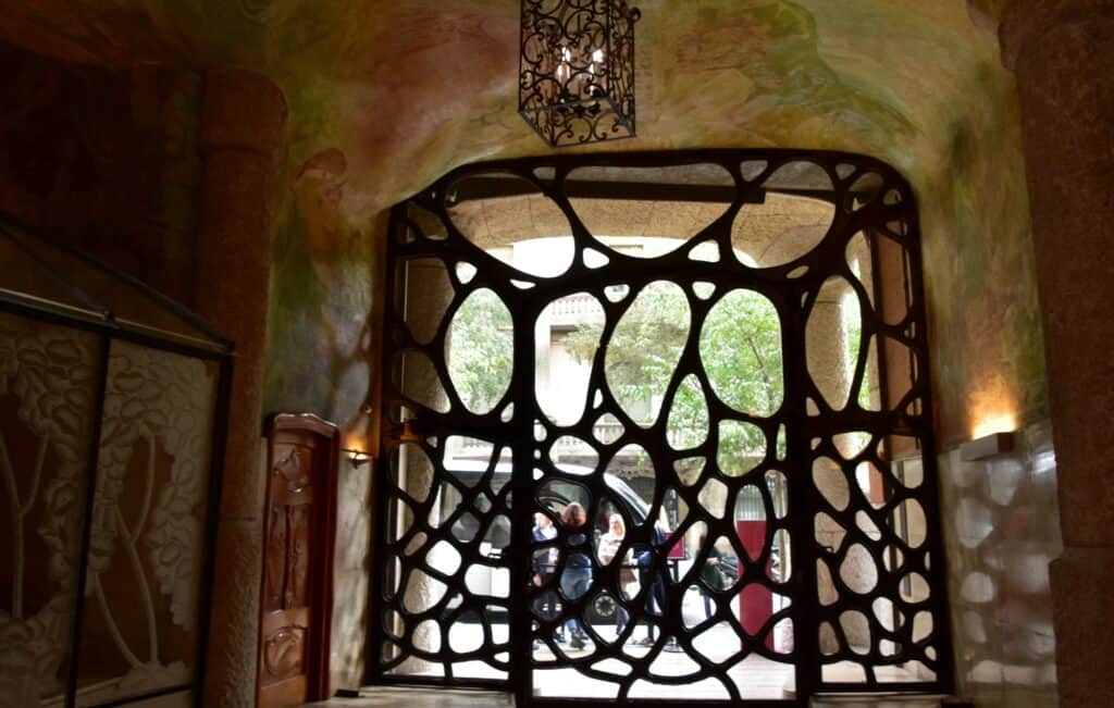 Organic iron entrance gate of Casa Milà seen from inside the painted hallway, irregular glazed openings like cells framing the street beyond, beneath a hanging lantern