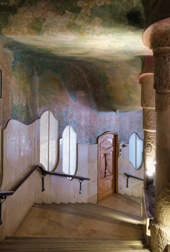 Grand stair to the main floor of Casa Milà, stone steps and a curving handrail rising beside oval windows and a carved timber door under a ceiling painted with mythological frescoes