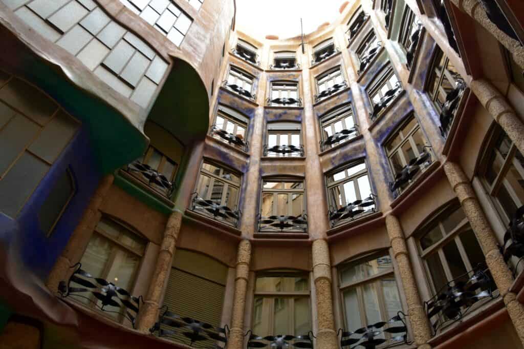 Looking up into a courtyard of Casa Milà, curving walls of windows and iron-railed balconies rising past painted soffits to an oval of sky