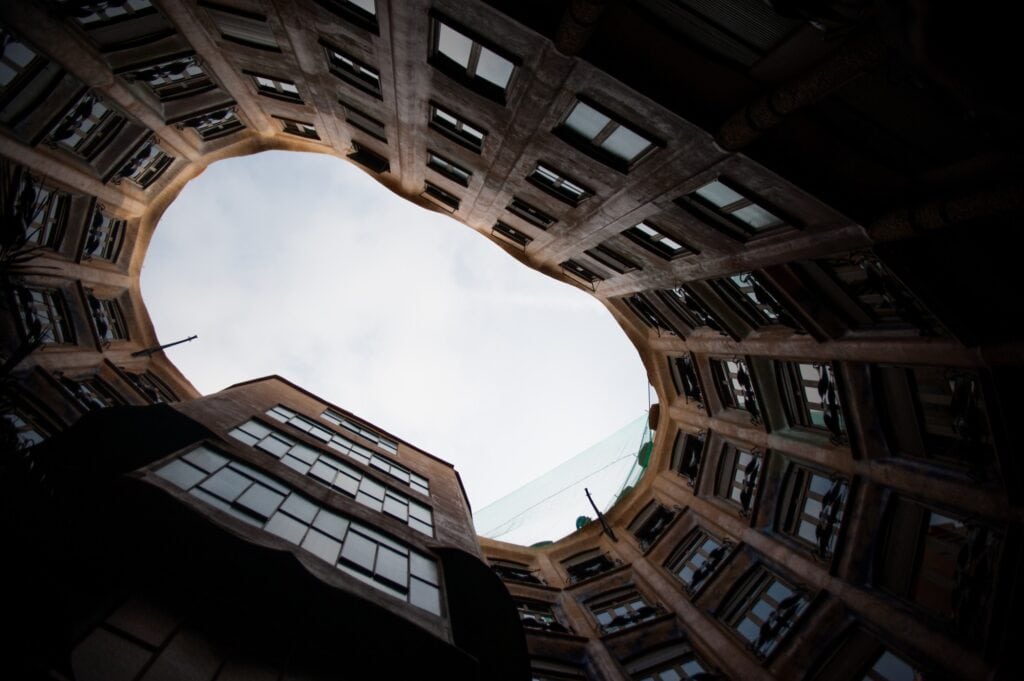 Dramatic upward view from the base of a La Pedrera courtyard, dark rings of windows and stone framing an irregular opening to a pale grey sky