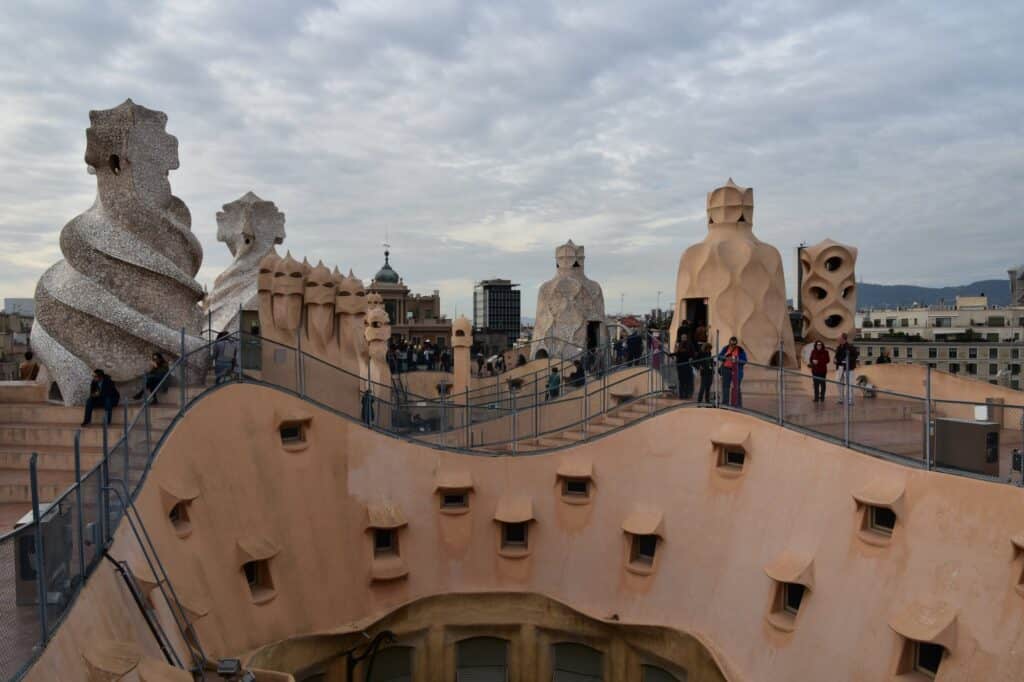 Undulating roof terrace of La Pedrera with visitors walking on its stepped paths among sculpted chimney stacks and stair towers, the Barcelona skyline beyond
