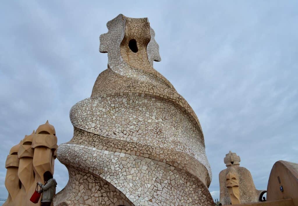 Spiralling stair tower on the roof of Casa Milà clad in white broken-marble mosaic and topped by a cross-shaped finial, with warrior chimneys on either side under a cloudy sky