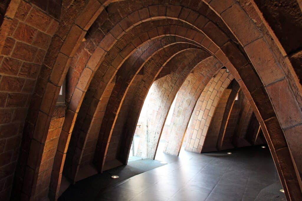 Sunlit brick catenary arches in the attic of La Pedrera, the parabolic ribs of thin Catalan vaulting curving over a dark floor towards small windows
