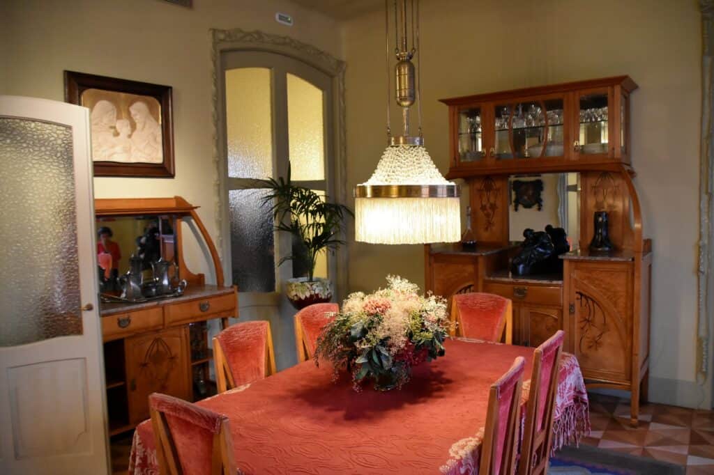 Dining room in the Pis de la Pedrera, a red-clothed table with flowers and upholstered chairs beneath a fringed pendant lamp, a carved Modernista sideboard and tall shuttered doors