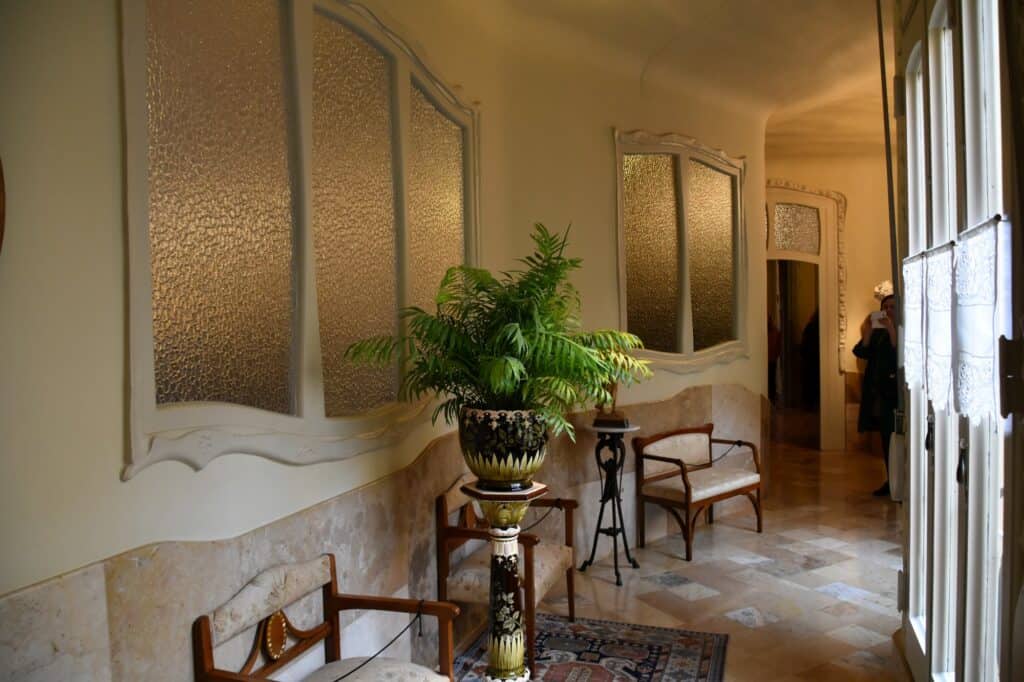 Hallway of the period apartment at Casa Milà with curved frosted-glass panels, a potted palm on a pedestal, Modernista chairs and patterned floor tiles in soft daylight