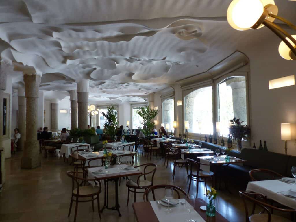 Café-restaurant at Casa Milà under a rippling white sculpted plaster ceiling, bentwood chairs and white-clothed tables between stone columns and tall windows