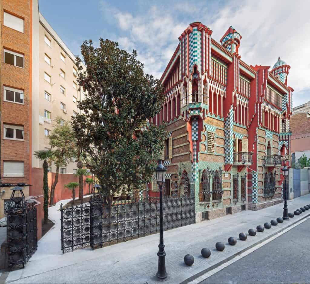 Casa Vicens on Carrer de les Carolines in Barcelona's Gràcia district, its red brick and green and white tiled façade topped by small corner turrets, behind a wrought-iron fence of palm-leaf panels and a leafy magnolia tree