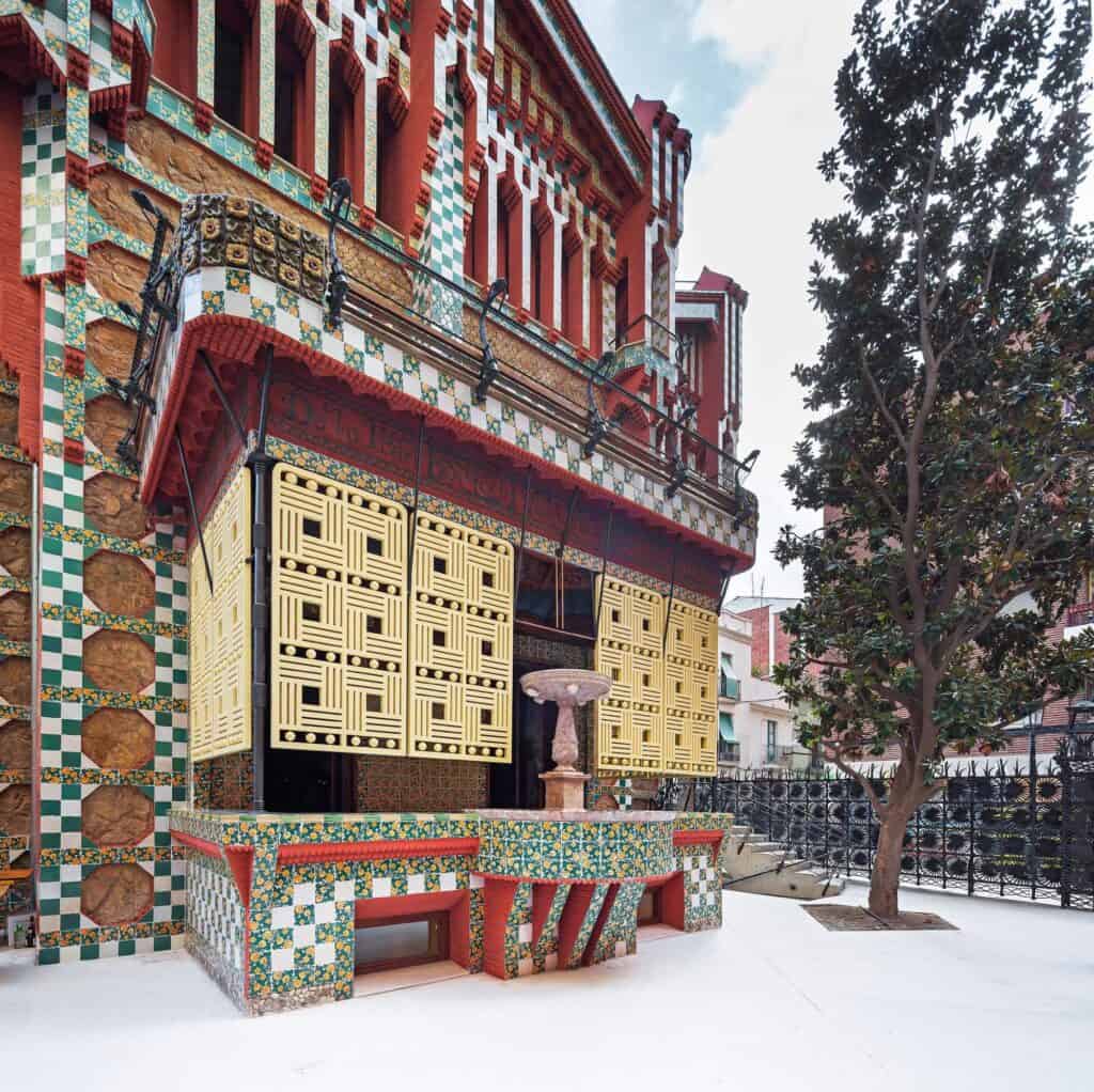 Garden façade of Casa Vicens with the enclosed porch projecting on red brackets, panels of yellow latticework shutters, a chequerboard of green and white tiles and a slim fountain on the porch roof, beside a tall cypress