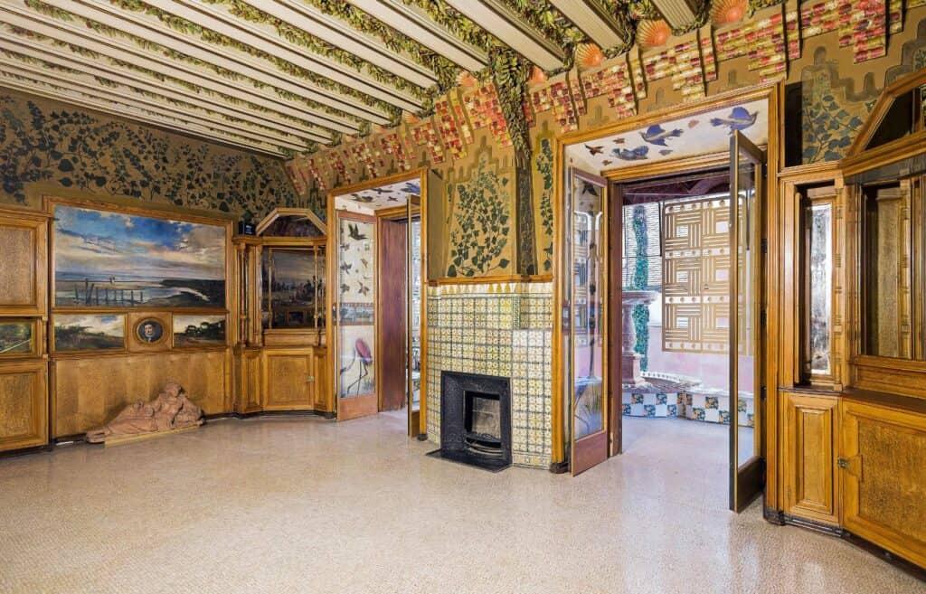 Restored dining room of Casa Vicens with built-in carved timber cabinets framing small oil paintings, a tiled fireplace, plant-motif sgraffito on the walls and painted ceramic panels between the ceiling beams