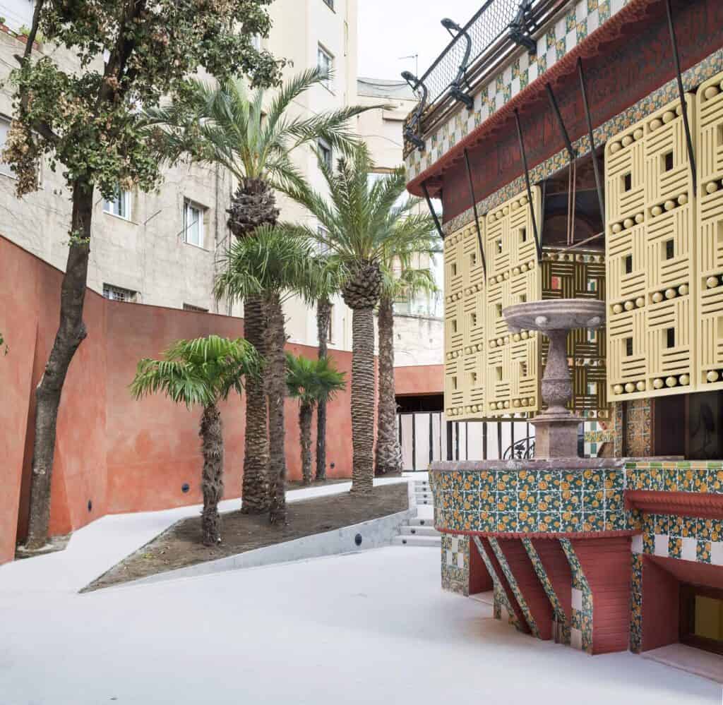 Corner of Casa Vicens seen from the garden, the porch with its fountain and latticework shutters above a red tiled base, fan palms and a large tree rising against a terracotta-red boundary wall