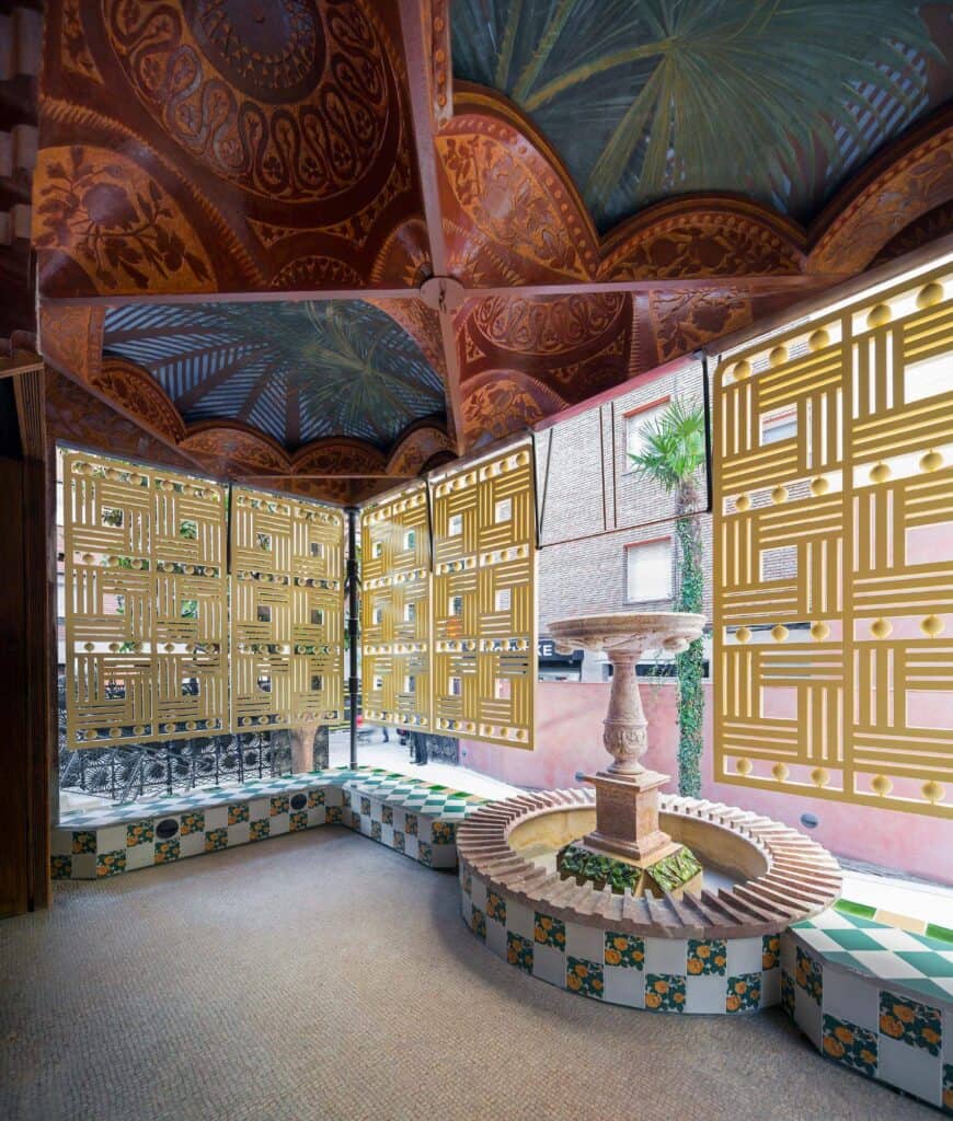 Enclosed porch of Casa Vicens with a recovered tiled fountain at its centre, walls of geometric yellow latticework and a ceiling painted with palm fronds and sky, framed by green and white tiles