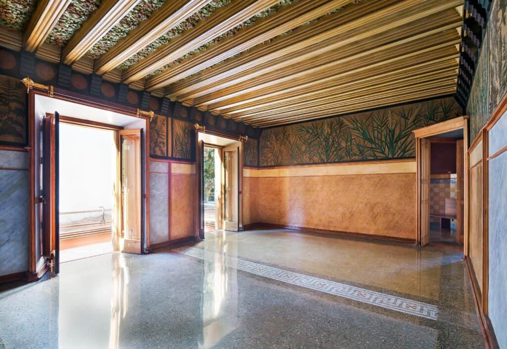 Long ground-floor room of Casa Vicens with gilded timber beams over painted ceramic panels, a band of ferns in sgraffito above a copper-toned dado, open timber doors to the garden and a polished terrazzo floor