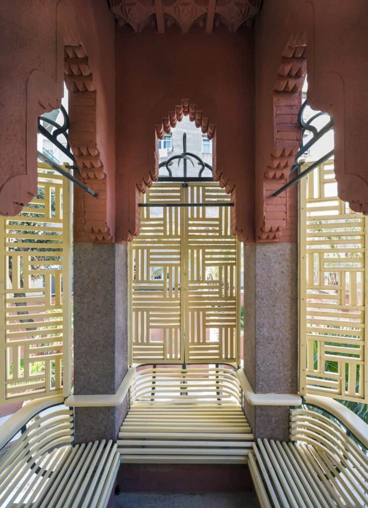 Window bay inside Casa Vicens with a cushioned bench under a carved terracotta arch of hanging mocárabes, framed by green and white tiles and screened by yellow geometric latticework shutters