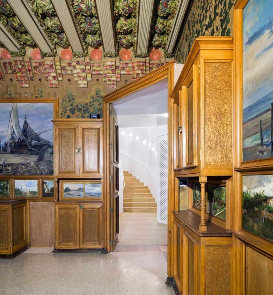 Corner of the Casa Vicens dining room with carved timber display cabinets and oil paintings of ships and landscapes, opening to the new white staircase of the museum beyond