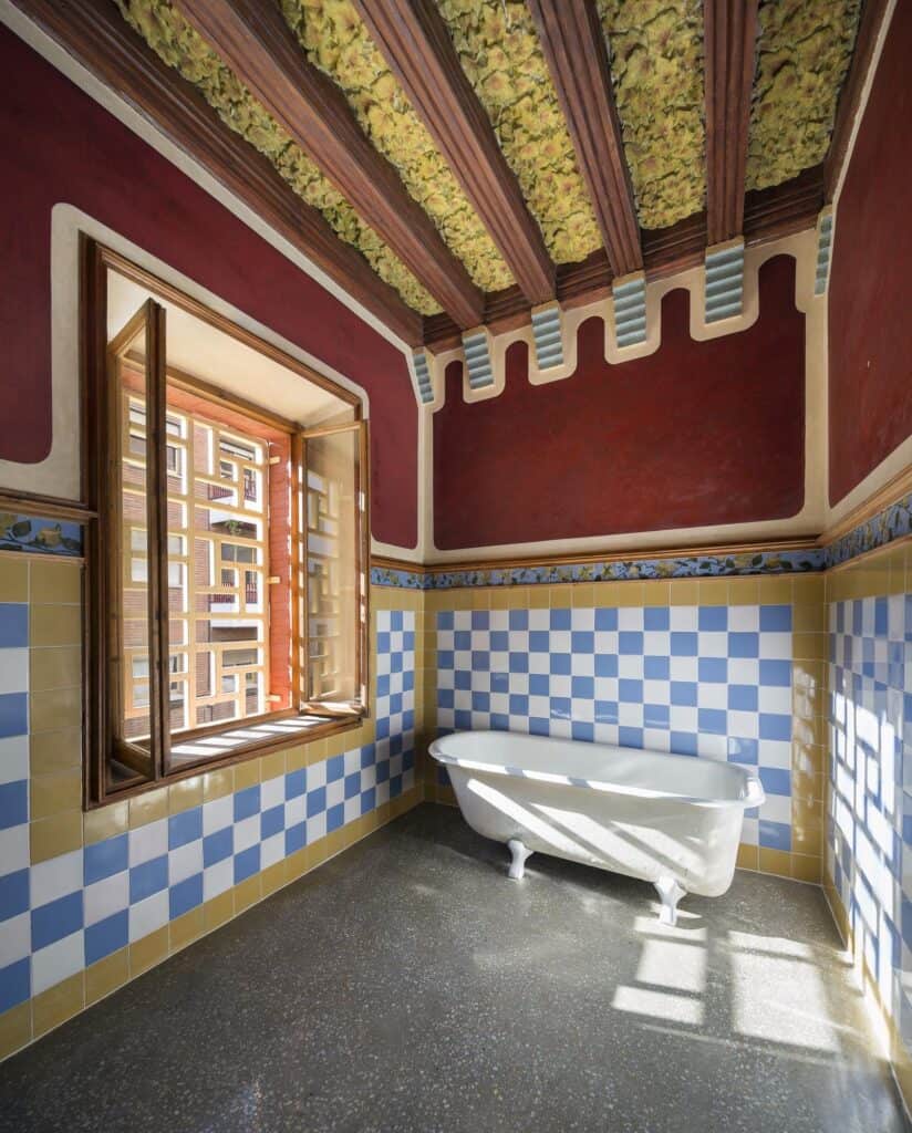 Restored bathroom at Casa Vicens with a white clawfoot bathtub on dark terrazzo, walls of blue and white chequered tiles under a deep red band, a timber window and a beamed ceiling painted with leaves