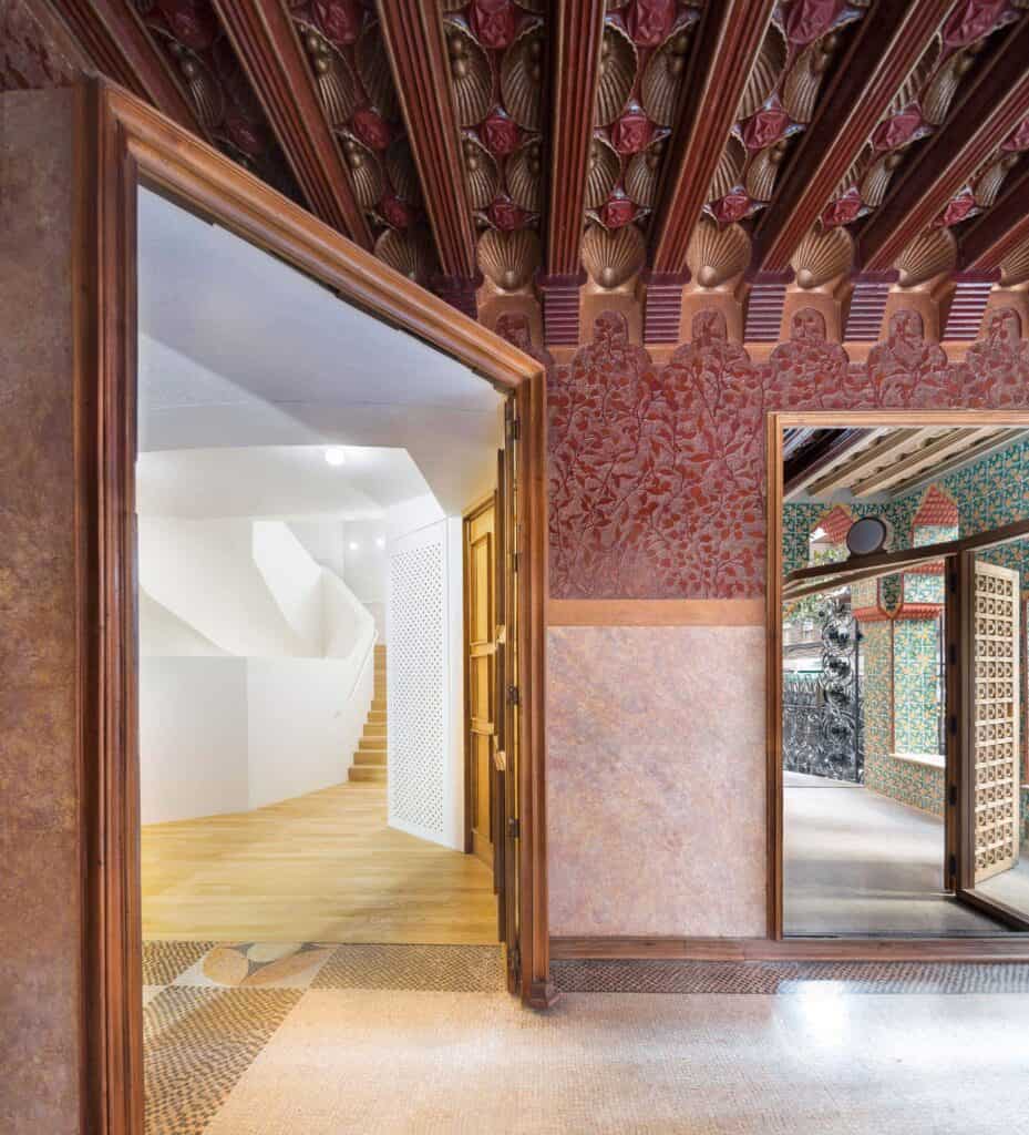 Doorway between the Gaudí house and the new museum stair at Casa Vicens, a red sgraffito wall of foliage beneath carved red and gold beams and a pink marbled dado, with a white stair glimpsed beyond