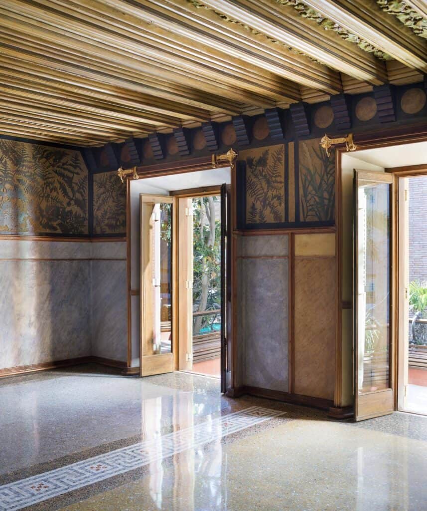 First-floor room of Casa Vicens with a gilded beamed ceiling, walls painted with dark leafy sgraffito above a grey marbled dado, tall timber doors open to a balcony and a terrazzo floor with a patterned border