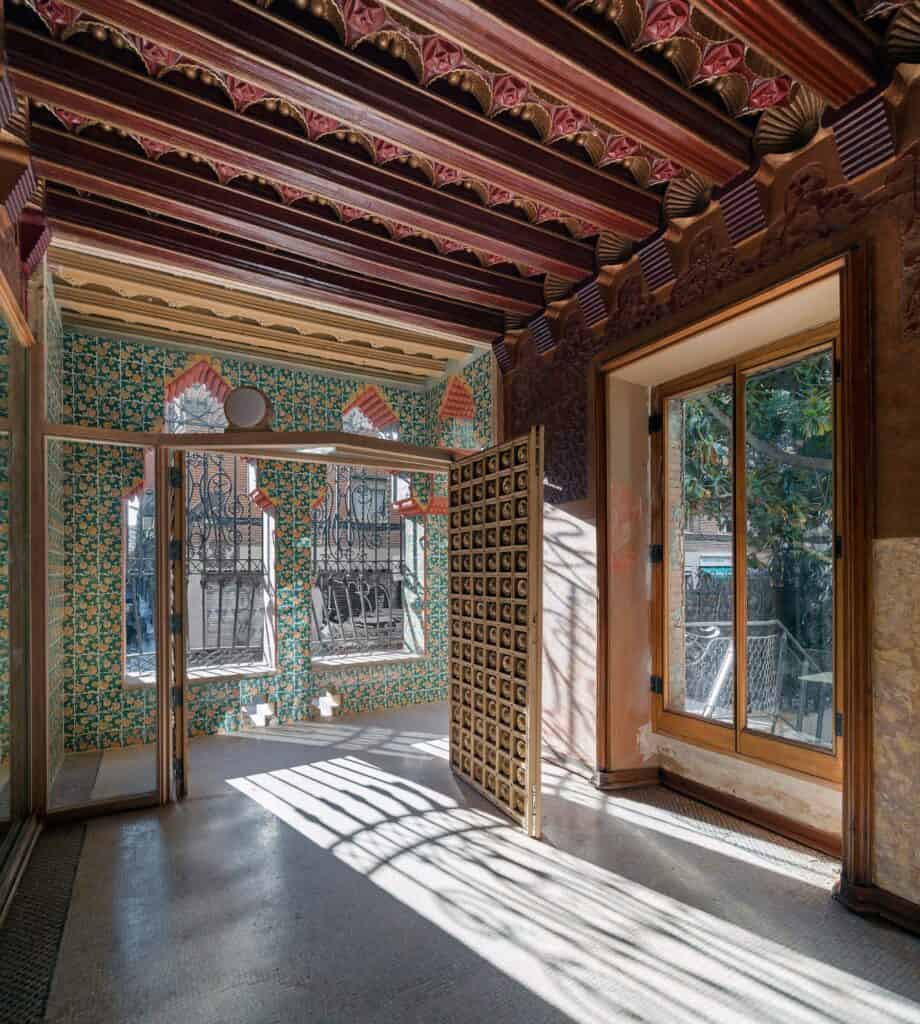 Room opening onto the enclosed porch at Casa Vicens, sunlight casting lattice shadows across the floor beneath a ceiling of carved and painted red and gold beams, green tiled walls and screened windows beyond
