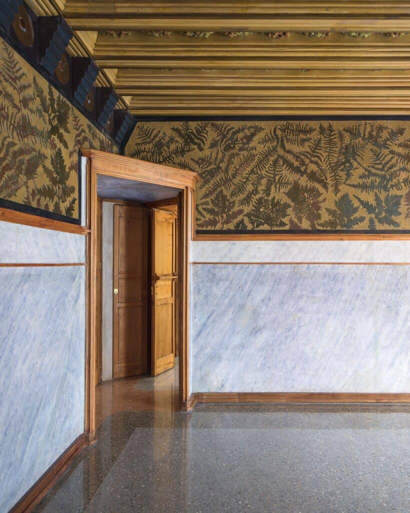 Restored bedroom at Casa Vicens with walls of dark green fern sgraffito above a pale grey marbled dado, a timber door with carved panels and a gilded beamed ceiling over a terrazzo floor