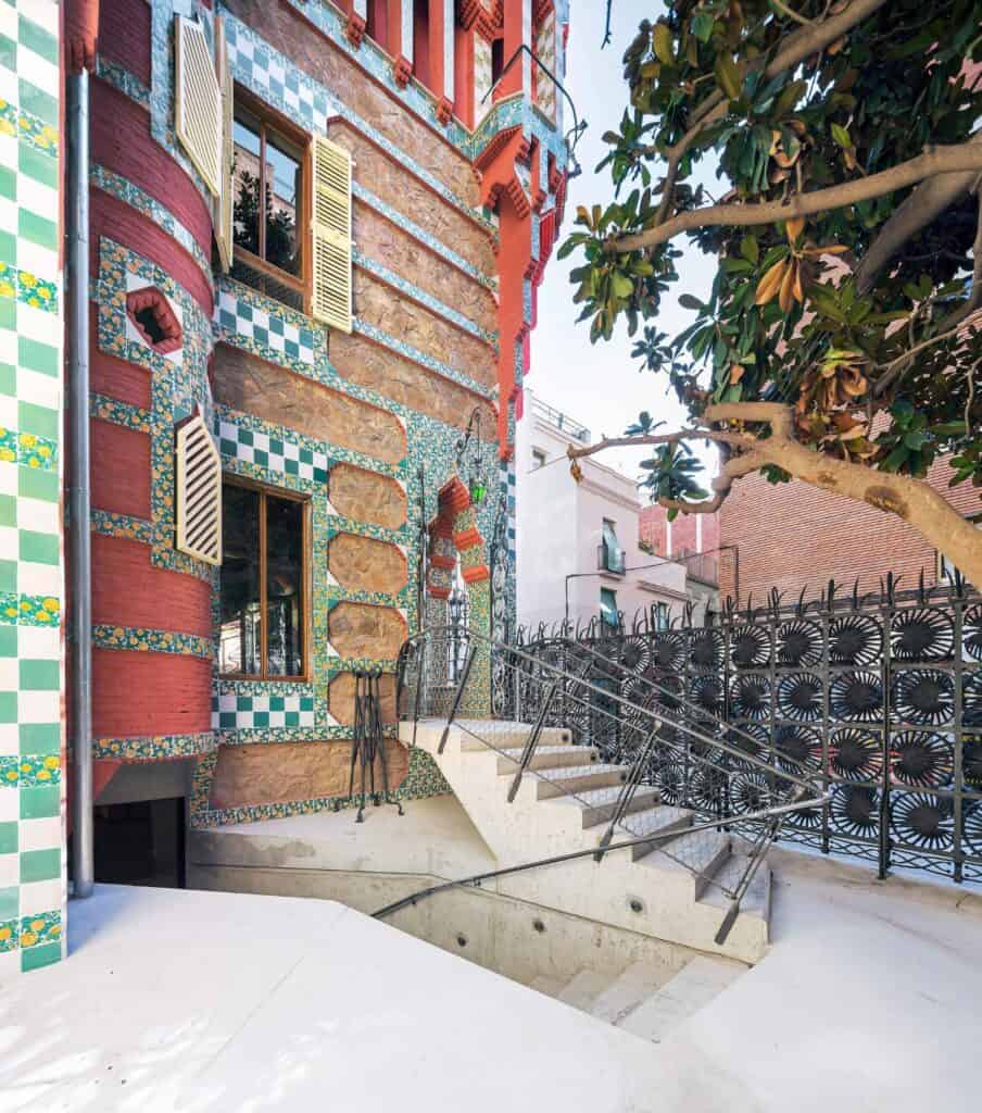 Side of Casa Vicens with a turret of red brick and green and white tiles, a new stone stair descending to the garden level beside the iron palm-leaf fence and an overhanging tree