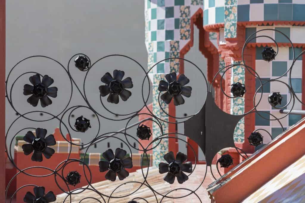 Wrought-iron railing of interlocking circles studded with black cast-iron flowers in front of a red brick turret and chequered green, white and pink ceramic tiles at Casa Vicens