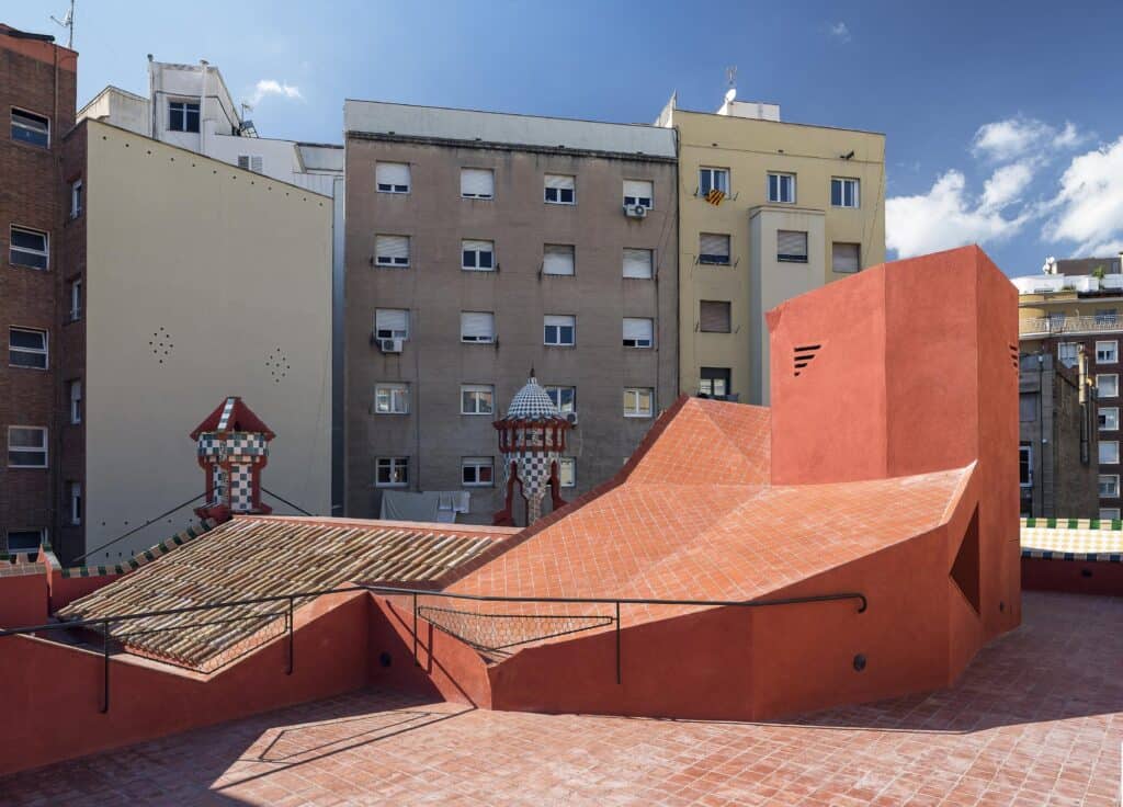 Restored rooftop of Casa Vicens with pitched tile roofs and two small tiled turrets beside an angular new terracotta-red volume housing the top of the stair, surrounded by apartment blocks under a blue sky