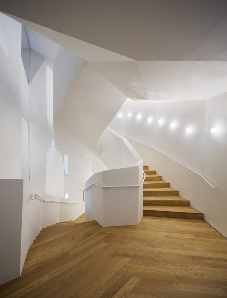 New white faceted staircase in the 1925 extension of Casa Vicens, oak treads rising past angled walls and a curving white balustrade lit by a row of small round lights