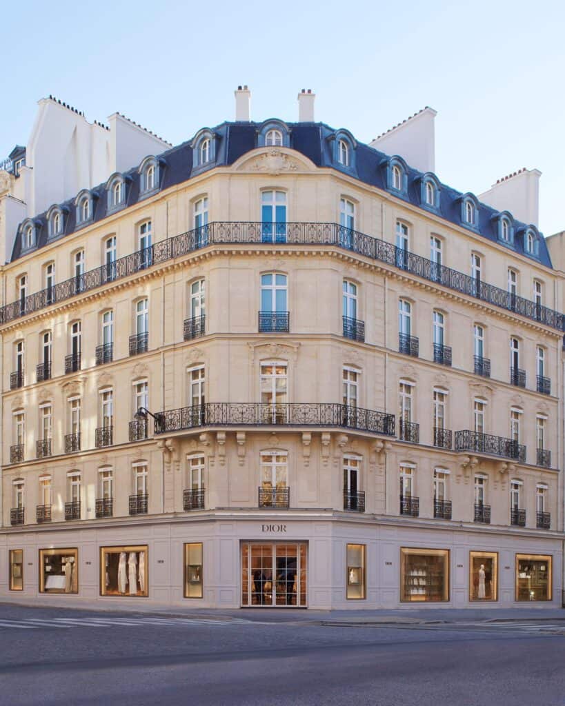 Corner facade of Dior 30 Montaigne on avenue Montaigne in Paris, a cream stone Haussmann-era building with wrought-iron balconies, a slate mansard roof with dormers and tall shop windows at street level