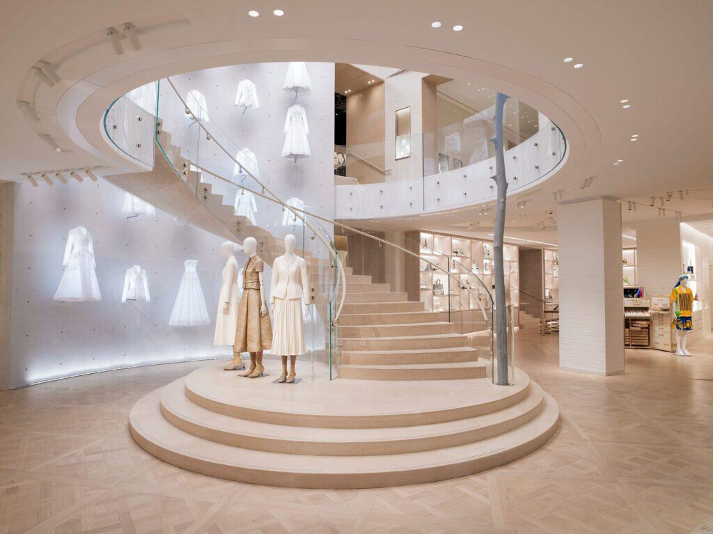 Triple-height rotunda at Dior 30 Montaigne, a sweeping white spiral staircase with a glass balustrade curving around two couture mannequins on a stepped circular plinth, white dresses floating on the wall behind