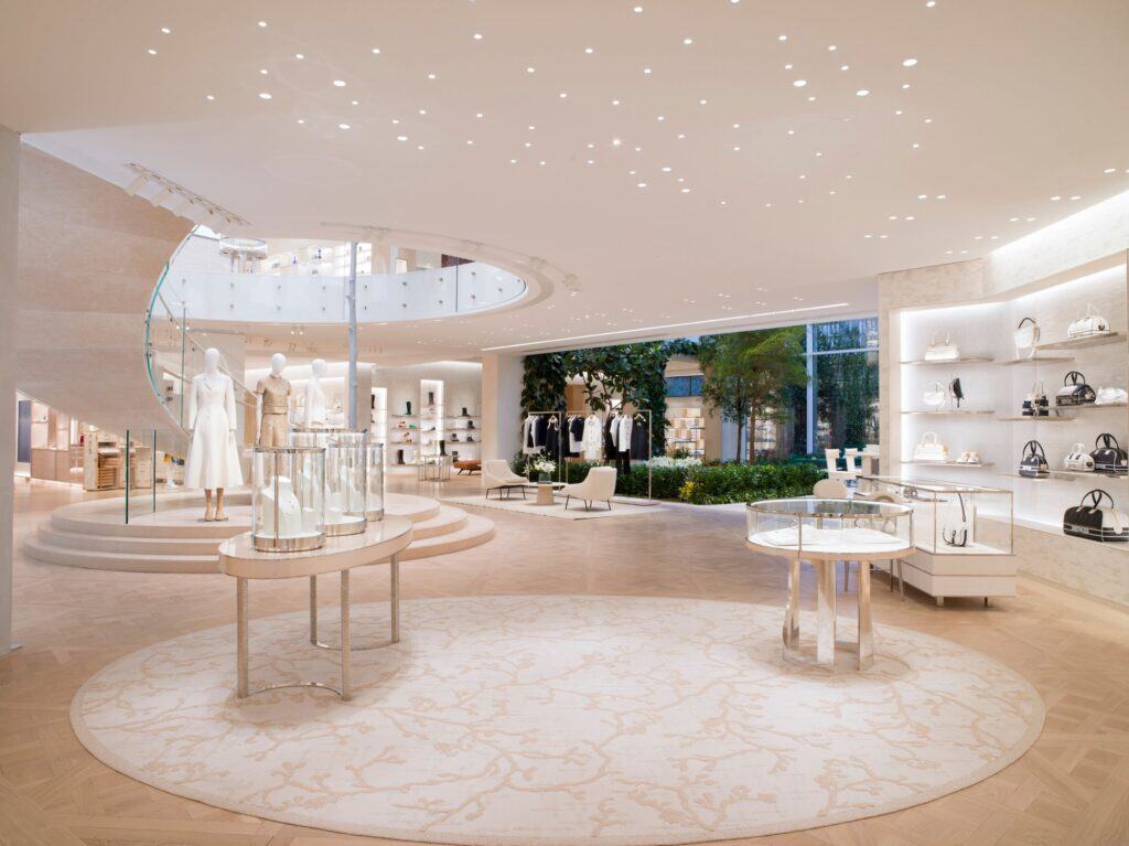 Boutique floor beneath the oculus of the rotunda at Dior 30 Montaigne, a round patterned rug, white display tables and shelves of handbags, with the palm-filled winter garden glimpsed beyond