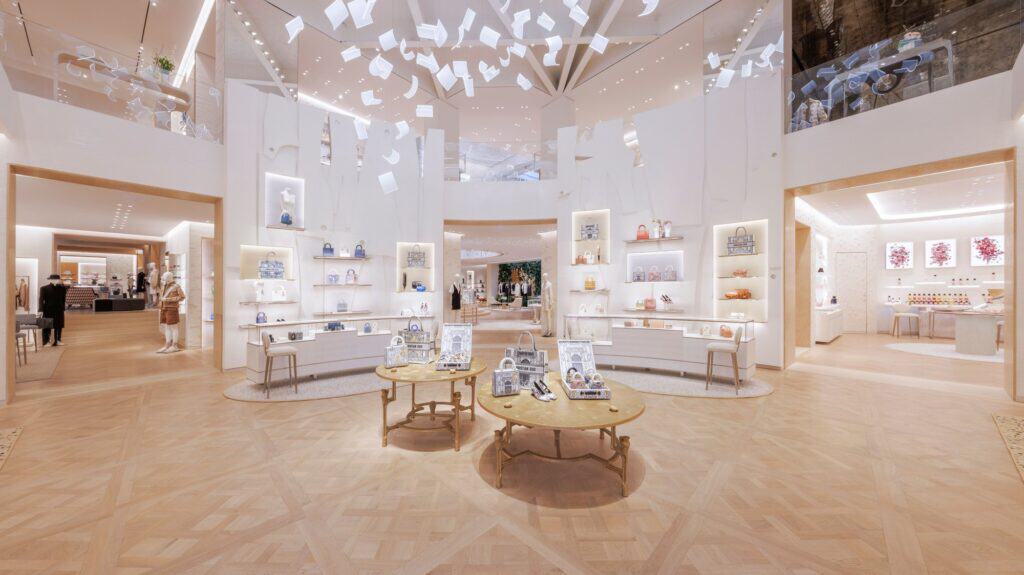 Wide view of a pale boutique hall at Dior 30 Montaigne, oak parquet, round tables of accessories and lit wall shelving of bags and shoes under a ceiling hung with white sculpted leaves