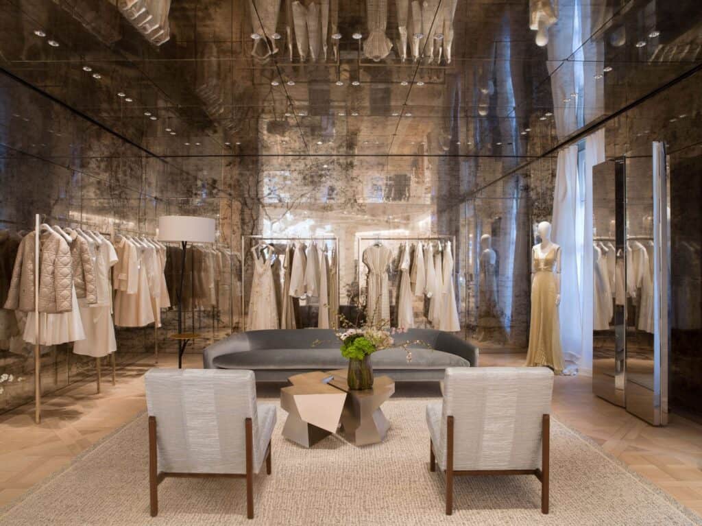 Ready-to-wear salon at Dior 30 Montaigne with mirrored walls and ceiling, rails of neutral couture garments, a grey sofa and two cream armchairs around a faceted metal coffee table
