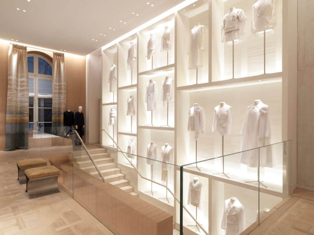Staircase at Dior 30 Montaigne climbing beside a wall of lit niches displaying white muslin toiles of Dior jackets and dresses, a tall arched window with sheer curtains on the left