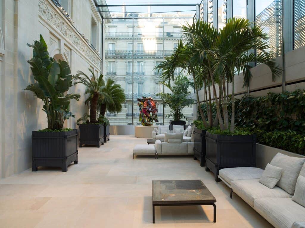 Glass-roofed winter garden at Dior 30 Montaigne with tall palms and leafy plants in black planters, low cream sofas and a dark coffee table on a pale stone floor between stone facades